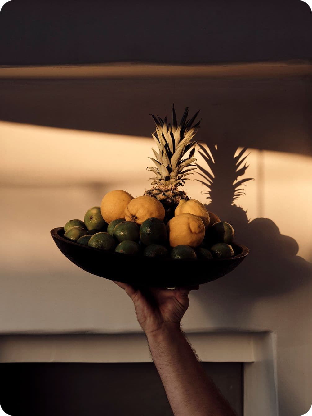 A hand holds a bowl of lemons, limes, and a pineapple, casting a shadow on a wall in warm, low lighting.