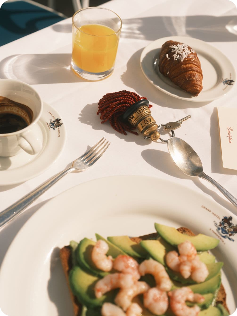 Plate with avocado and shrimp toast, a croissant, coffee, orange juice, and a hotel key on a white tablecloth in sunlight.