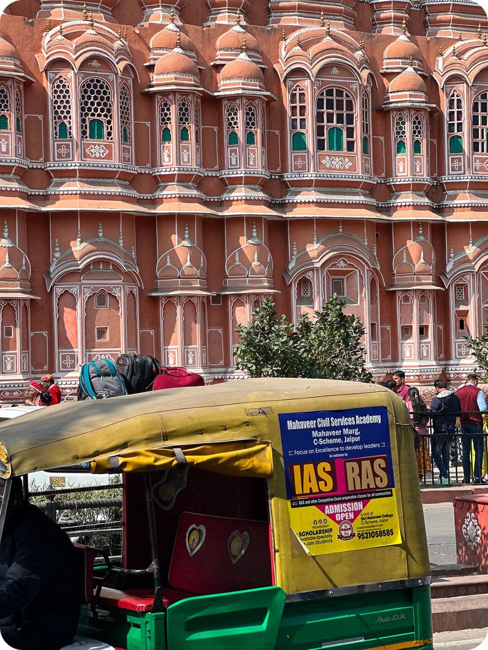 Auto-rickshaw in front of the ornate pink facade of Hawa Mahal, Jaipur, with people and trees in the background.