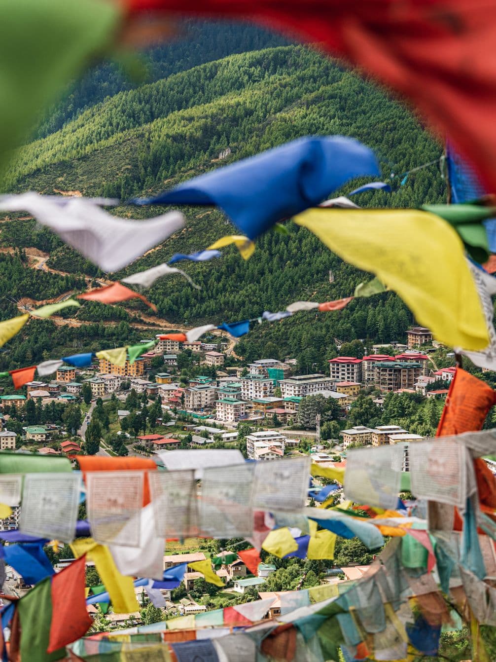 Colorful prayer flags fluttering over a hillside town surrounded by lush green mountains, with a variety of buildings visible in the background.