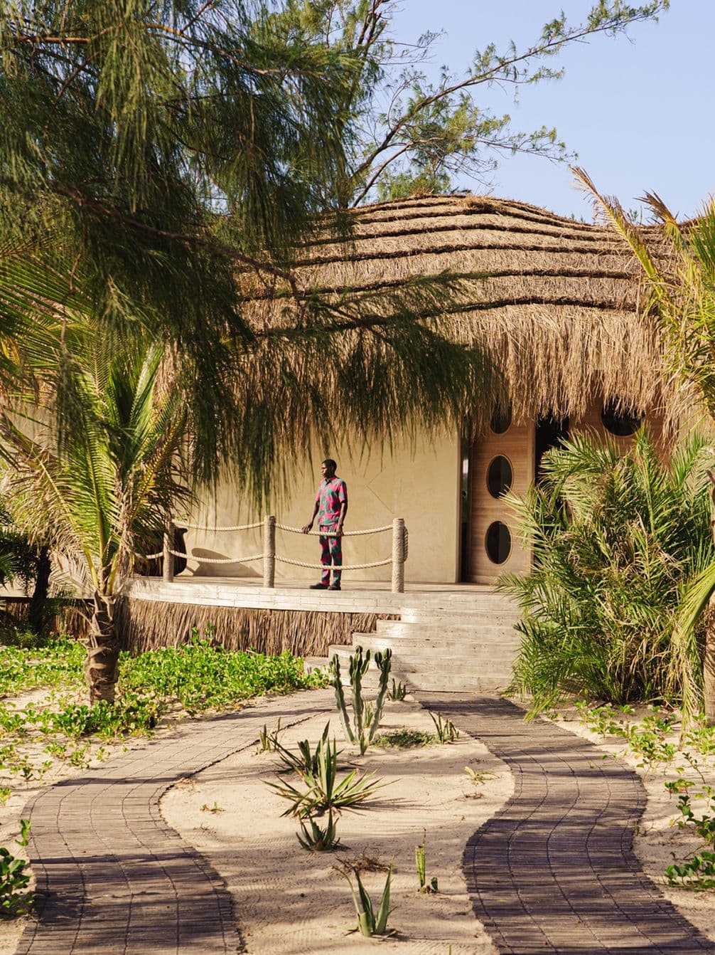 Thatched-roof bungalow surrounded by tropical plants, with a person standing at the entrance, on a sunny day.