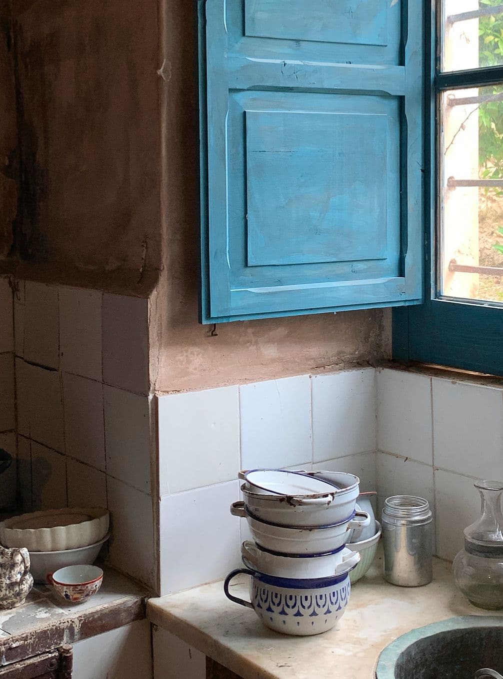 A rustic kitchen scene with stacked enamel mugs, a glass jar, and a ceramic bowl on a tiled counter. A blue shuttered window lets in natural light.