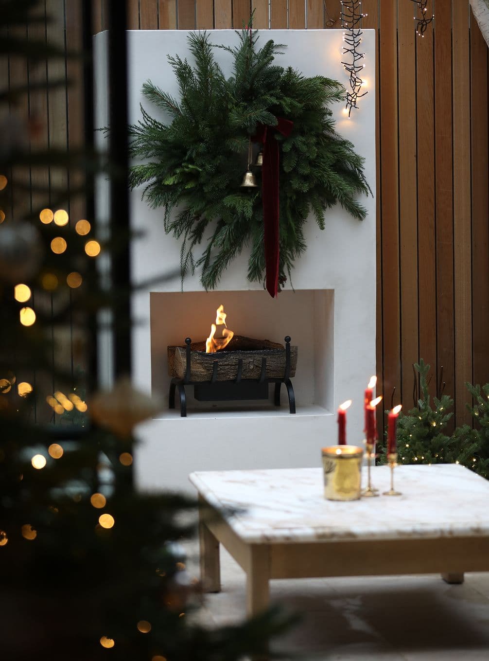 Cozy outdoor fireplace with a festive evergreen wreath, red ribbon, and glowing candles on a wooden table. Christmas lights twinkle nearby.
