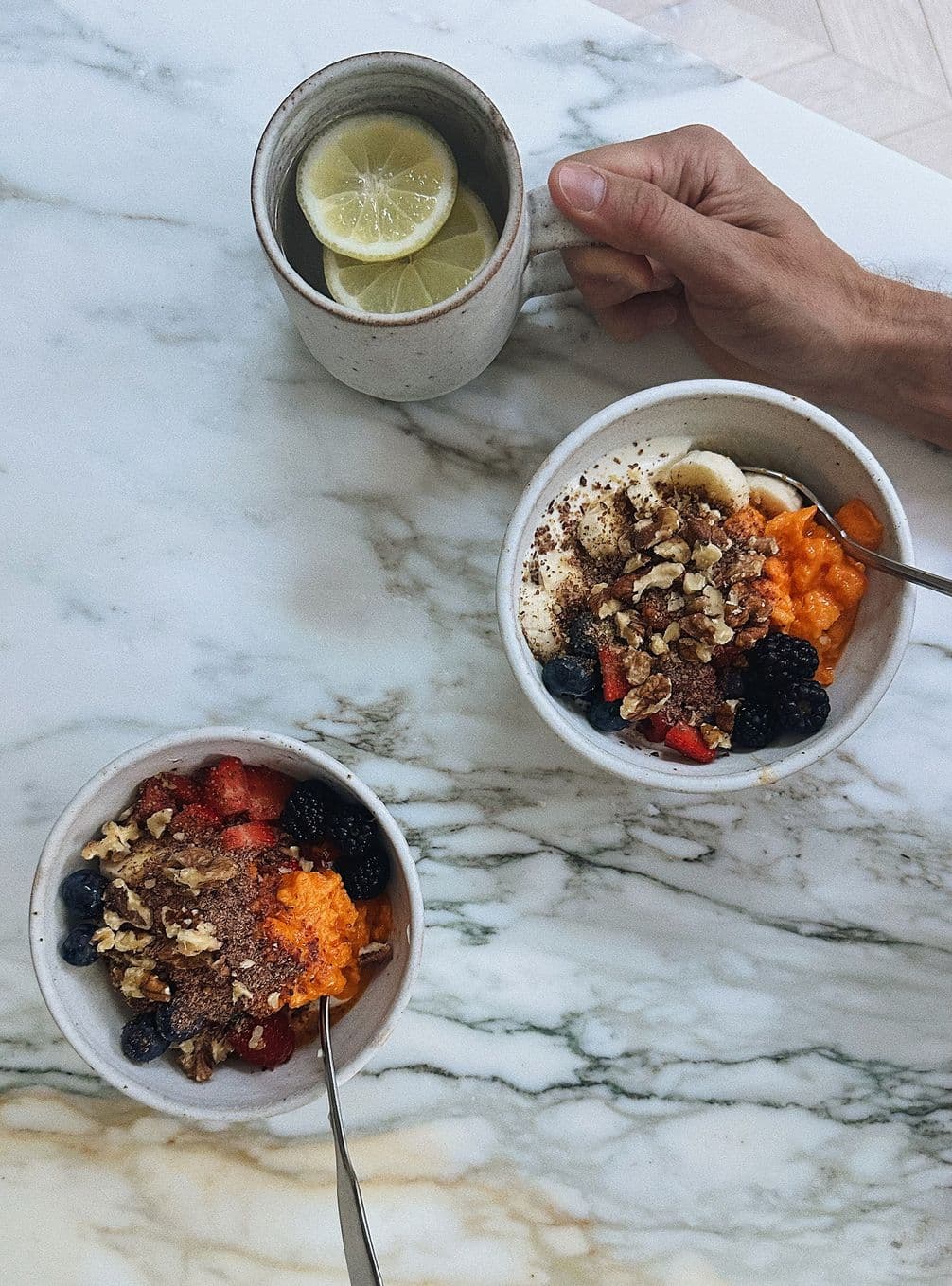 Two bowls of fruity granola with berries and nuts on a marble surface, next to a lemon-infused drink held by a hand.