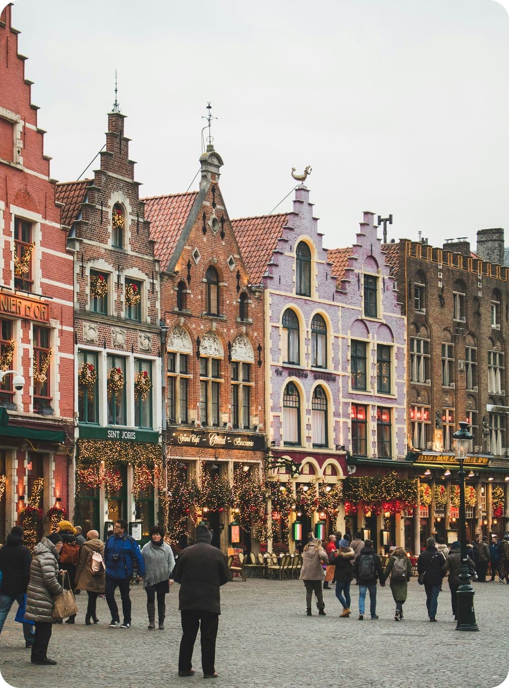 Colorful historic buildings with stepped gables, adorned with festive lights, line a cobblestone street bustling with people.