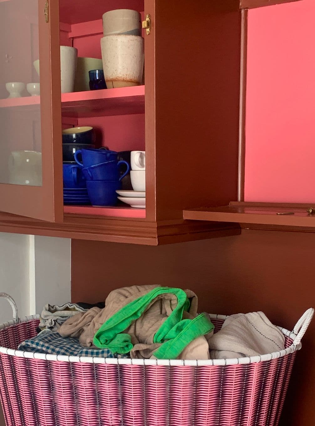 A pink basket with clothes sits below a brown cabinet displaying blue and white dishes and cups against a pink interior backdrop.