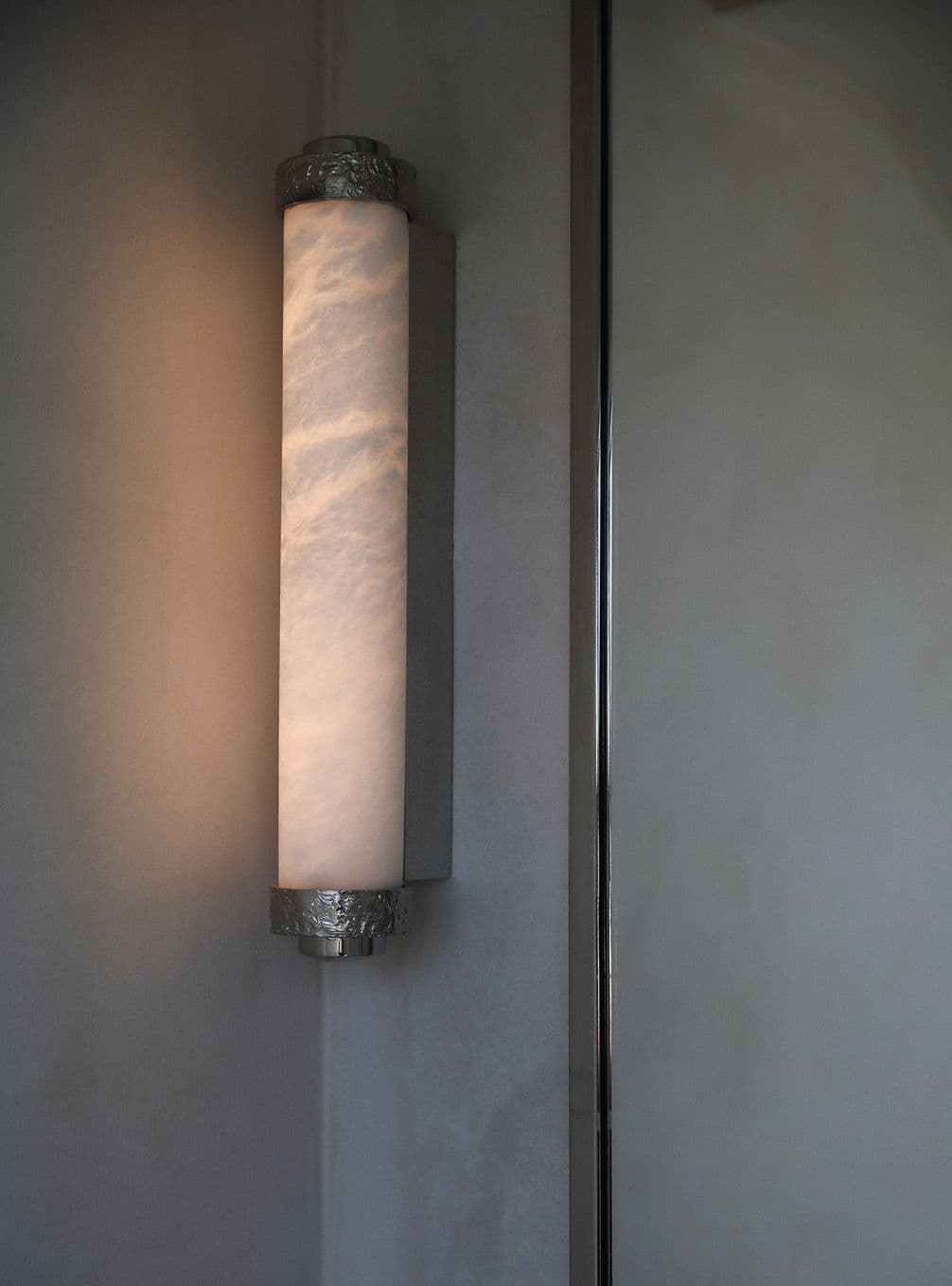 A vertical, cylindrical wall sconce emits soft white light, mounted on a smooth gray wall with metal accents at the top and bottom.
