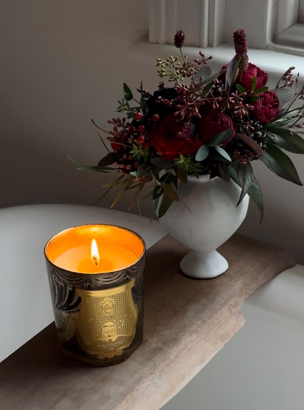 A lit candle in a decorative holder beside a vase with a bouquet of red and dark flowers on a wooden surface near a window.