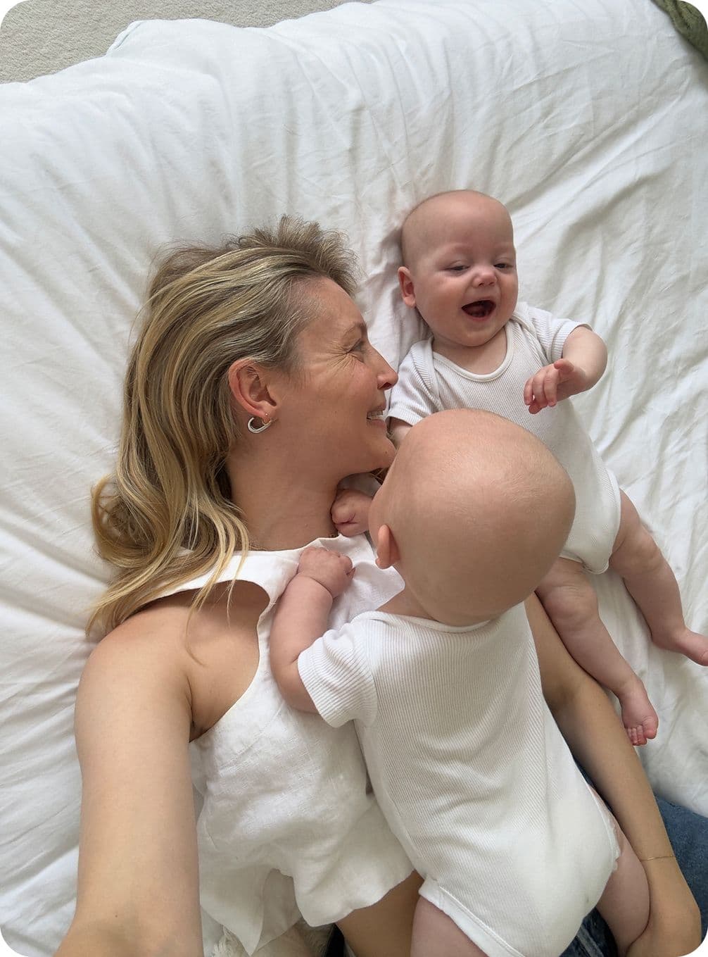 A woman lies on a bed, smiling at two babies in white onesies. One baby is lying on her chest, and the other is laughing beside her.