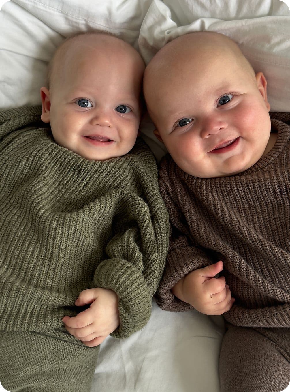 Two smiling babies in knitted sweaters, one in green and the other in brown, lying on a white blanket, facing the camera.