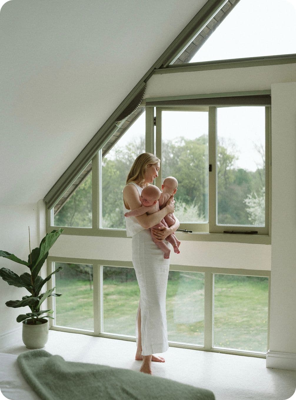 A woman in a white dress stands by large windows, holding two babies. A potted plant is nearby, and greenery is visible outside.