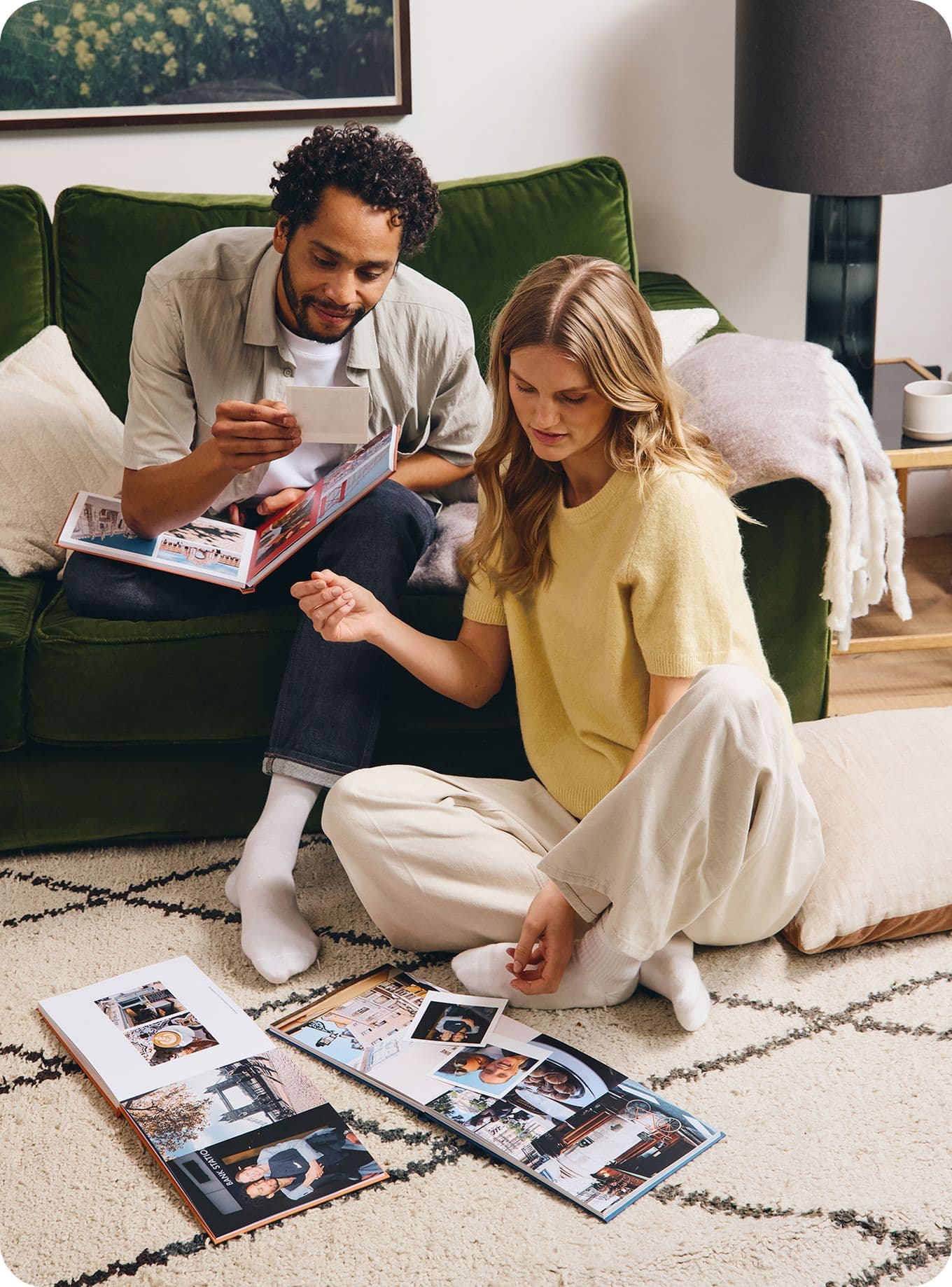 A man and woman sit on the floor, looking at photo albums and pictures in a cozy living room with a green sofa and a framed picture on the wall.