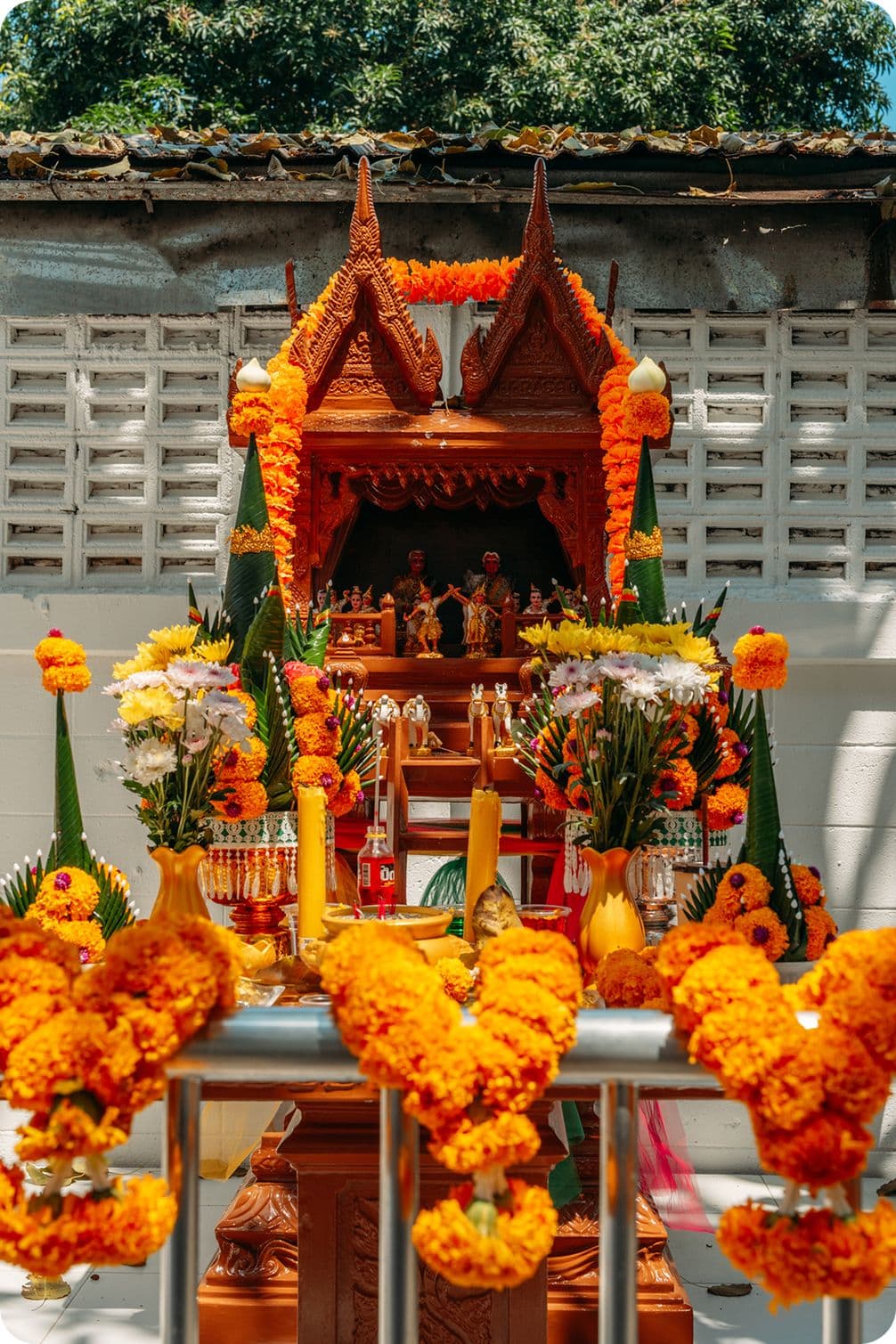 Ornate wooden Thai spirit house shrine adorned with bright orange marigold garlands, candles, and colorful floral offerings with miniature figurines.