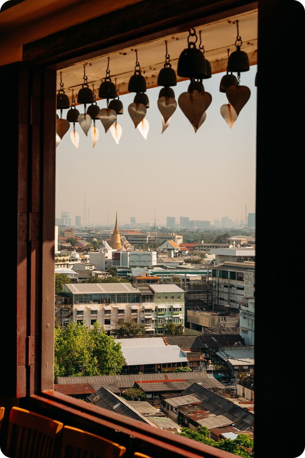 Urban skyline viewed through a wooden window, foreground of hanging temple bells with heart-shaped wind catchers and a distant golden spire.