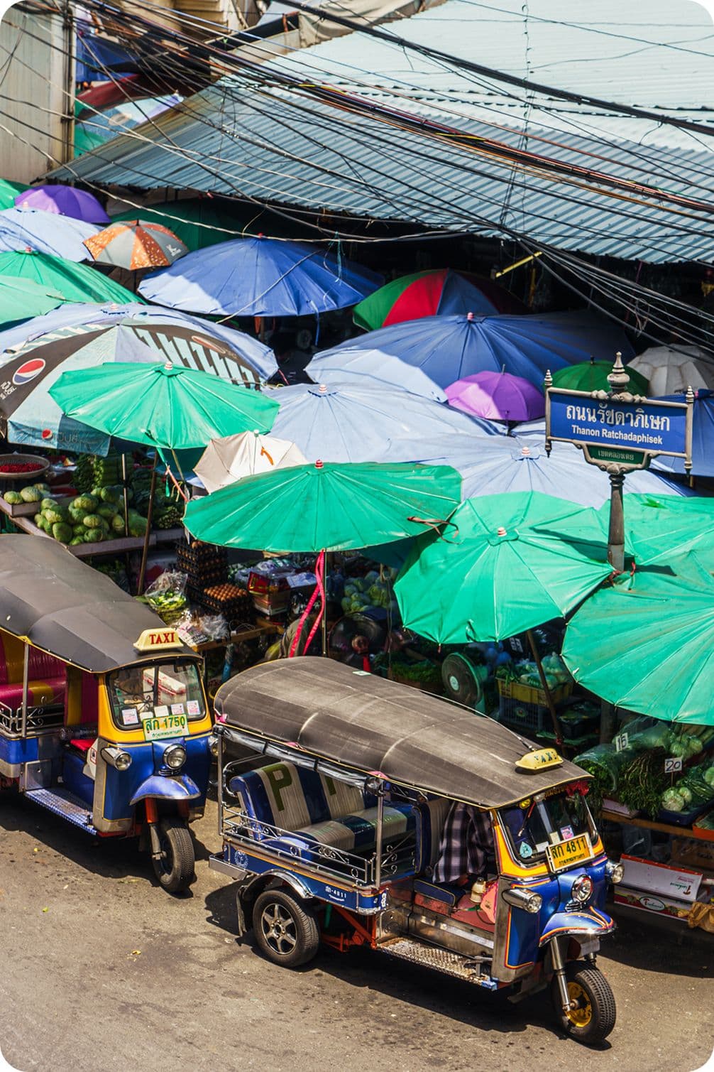 Two tuk-tuks parked beside a bustling Thai street market crowded with green and blue umbrellas and stalls of produce.