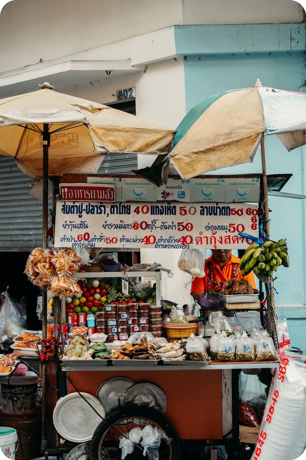 Thai street food cart under umbrellas, vendor selling fruits, prepared foods and jars, bunch of bananas and Thai price sign.