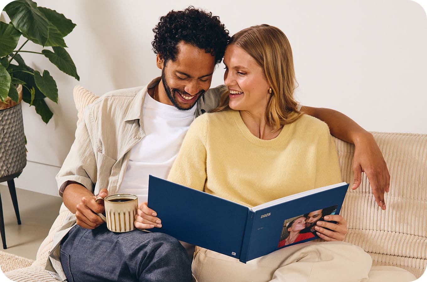 A smiling couple sits on a couch, looking at a photo album together. One holds a mug. A potted plant is in the background.