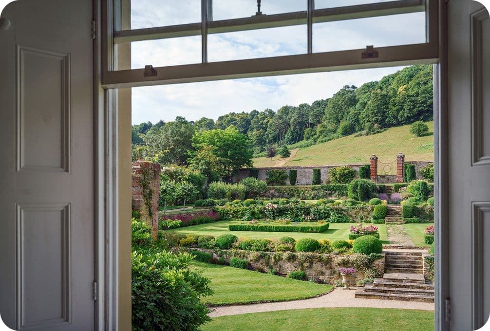 View through an open window of a landscaped garden with hedges, steps, and a hillside in the background under a partly cloudy sky.