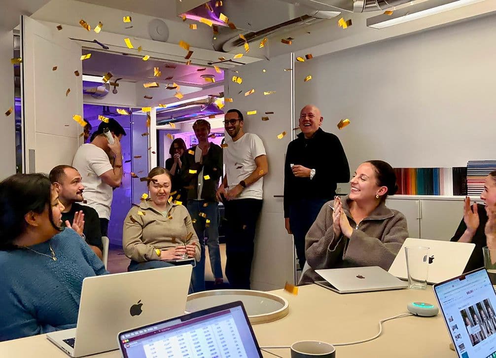 People in an office celebrate with confetti, while sitting around a table with laptops. Smiles and applause fill the room.