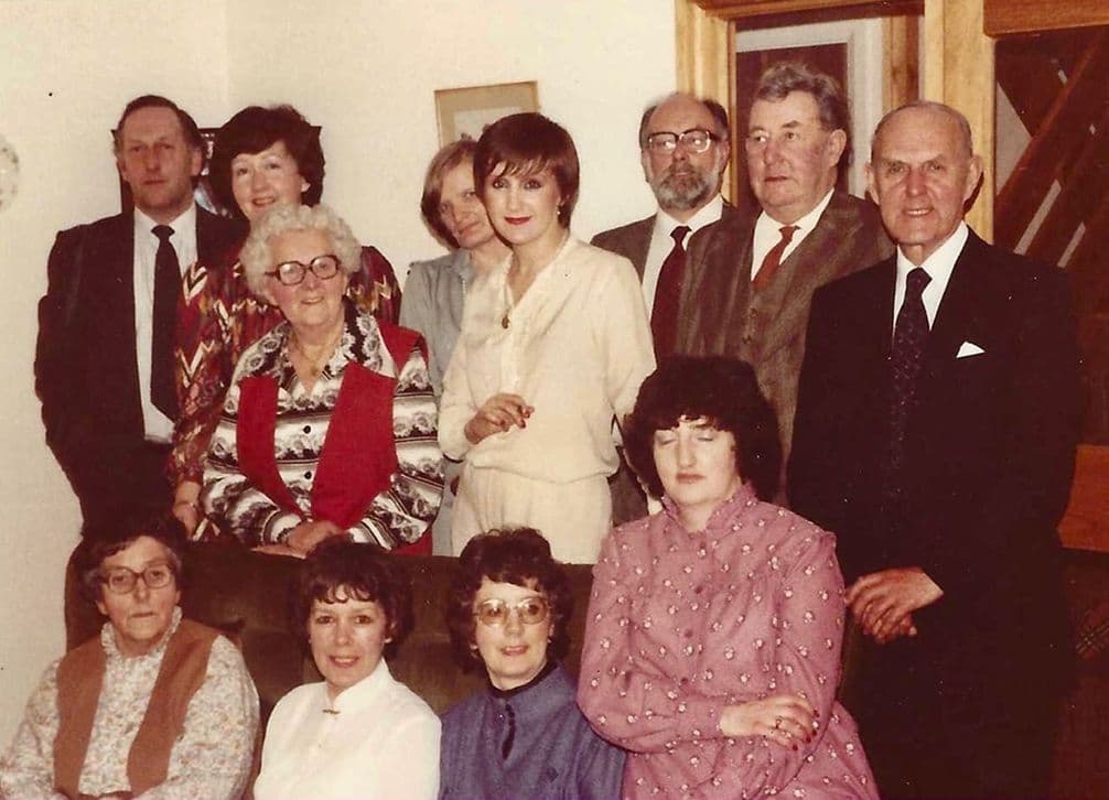 Group of thirteen people indoors, some seated, others standing, dressed in formal and semi-formal attire, posed for a portrait in a room.