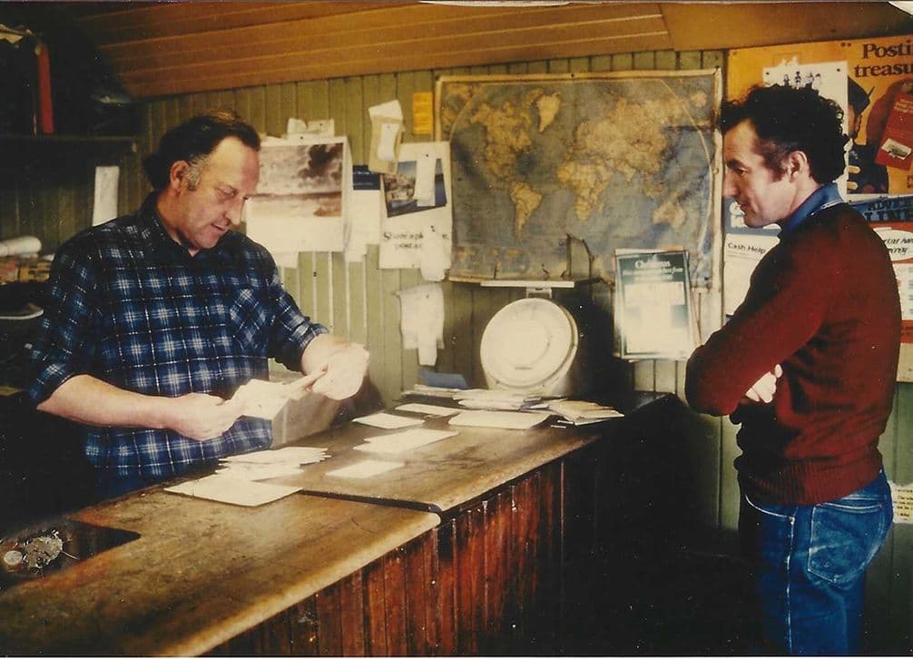 Two men in a rustic room with a world map on the wall; one sorts papers at a wooden counter while the other watches.