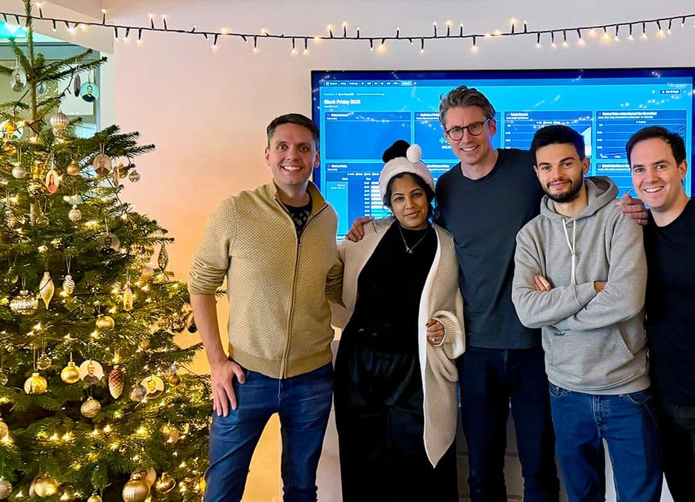 Five people posing next to a decorated Christmas tree and a large screen, with festive lights hanging above.