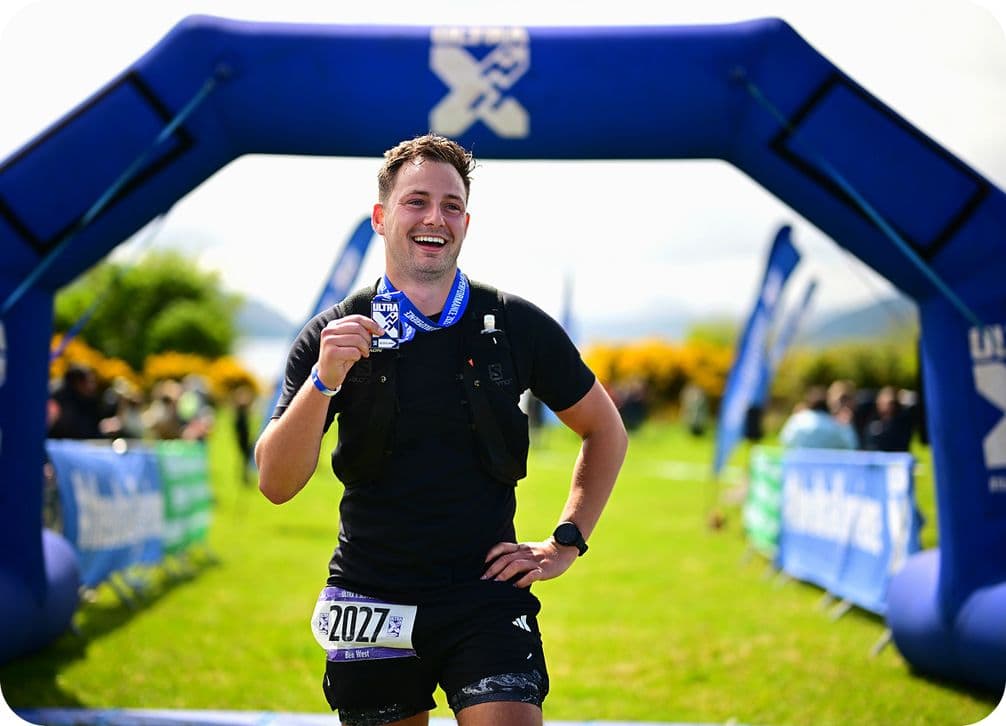 Runner smiling, holding a medal, stands under a blue finish line arch. He's wearing a black outfit with a race bib number 2027.