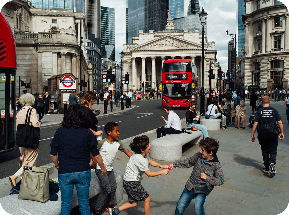 People walking and sitting near the Bank of England in London, with a red double-decker bus and surrounding historic buildings in view.
