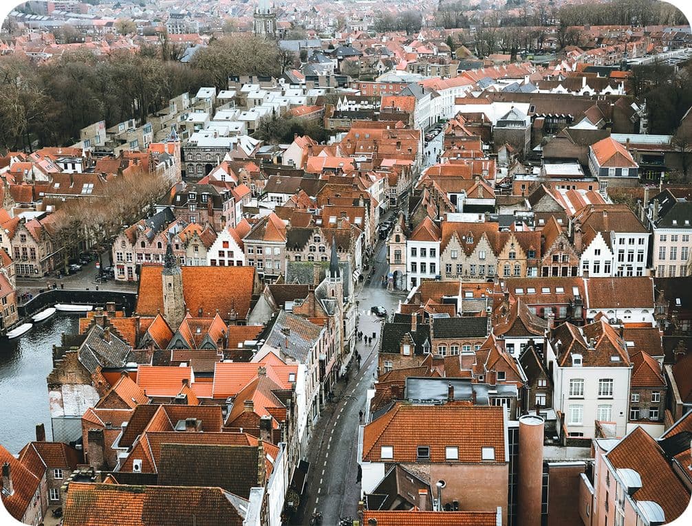 Aerial view of a historic European town with red-roofed buildings, narrow streets, and a canal, surrounded by trees and dense urban architecture.