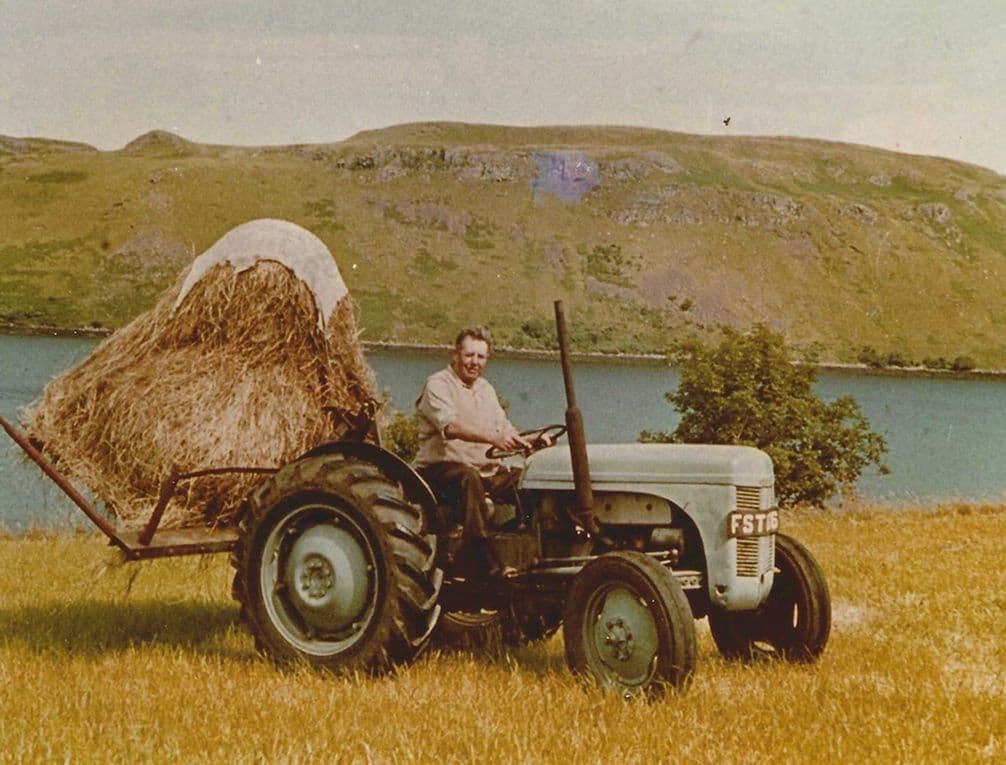 A person drives a tractor hauling hay in a field, with a lake and hills in the background.