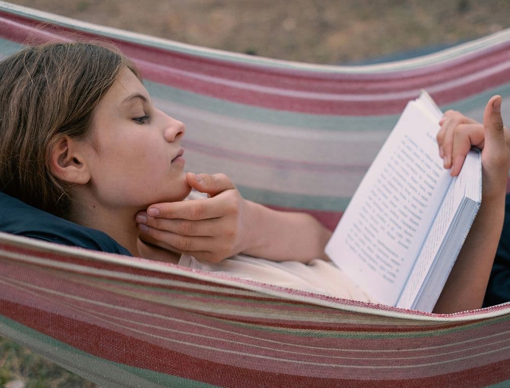 Person reclining in striped hammock reading a book, hand resting under chin.