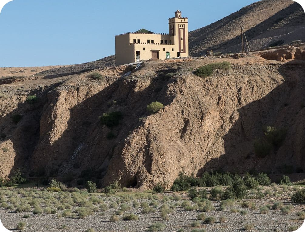A solitary building with a tower sits atop a rocky hill, overlooking a dry, sparse landscape under a clear blue sky.