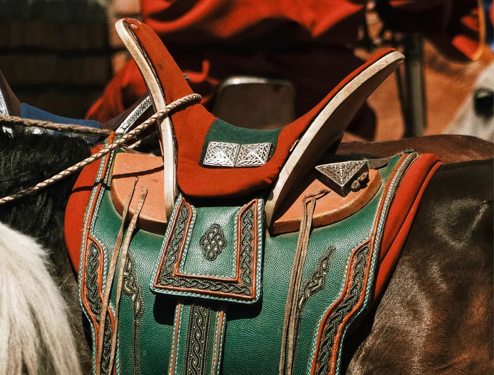 Ornate horse saddle with green and red leather, detailed stitching, and metal accents, mounted on a horse.