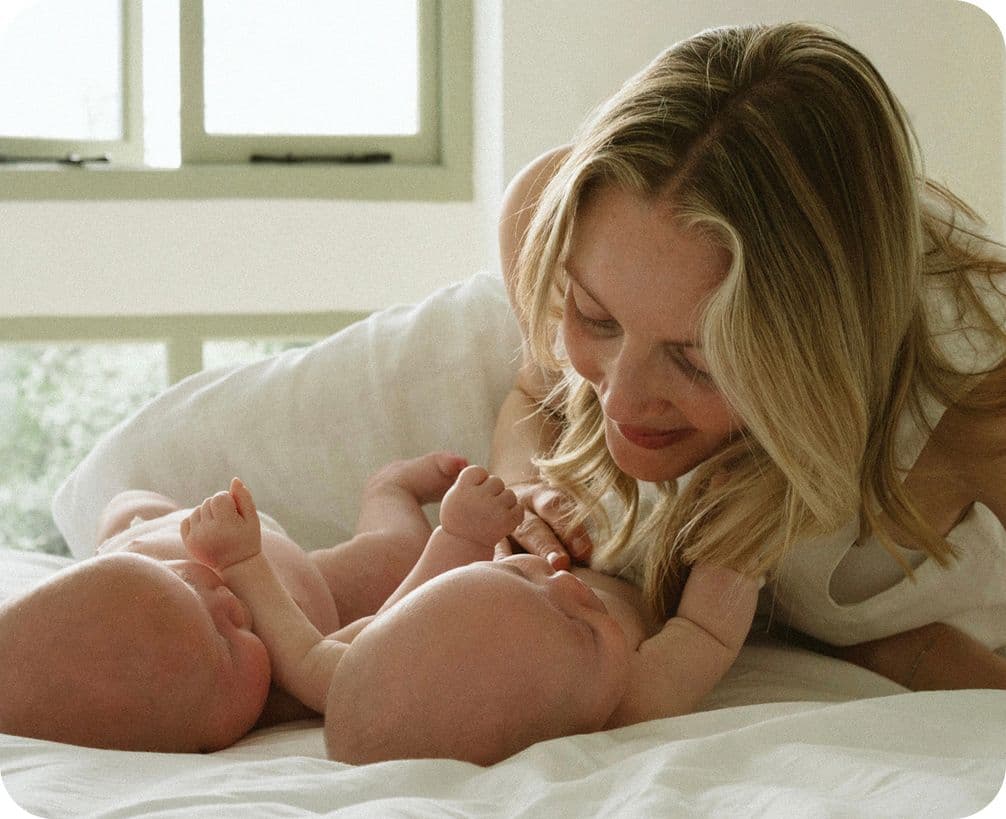 A woman smiles lovingly at two babies lying on a bed in a softly lit room with a window in the background.