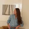 A woman in a blue shirt and jeans stands indoors, looking to the side, with a framed window photo and a wooden bowl in the background.
