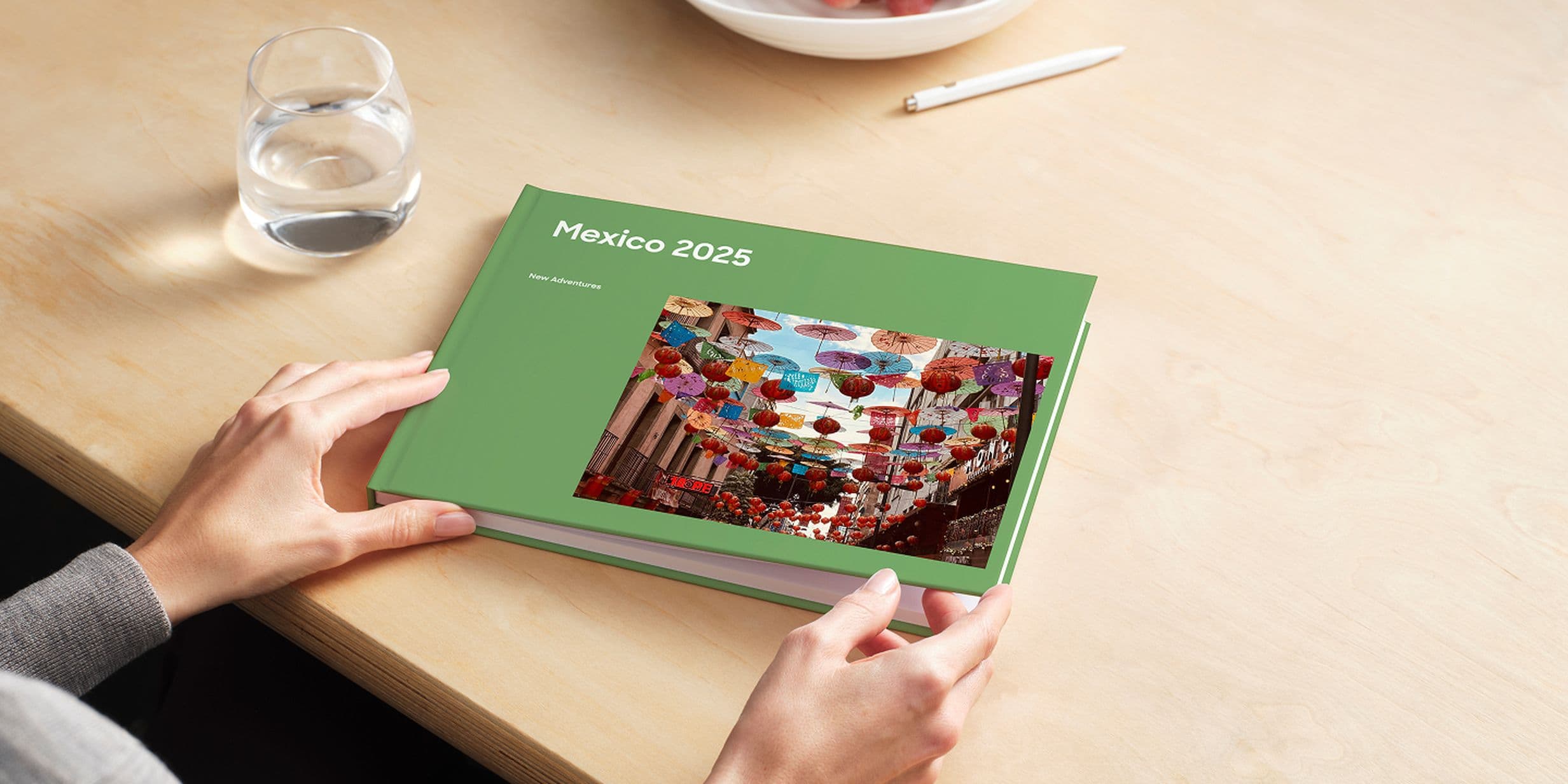 Person holding a green photo book titled "Mexico 2025" with a colorful market scene on the cover, on a wooden table with a glass of water.
