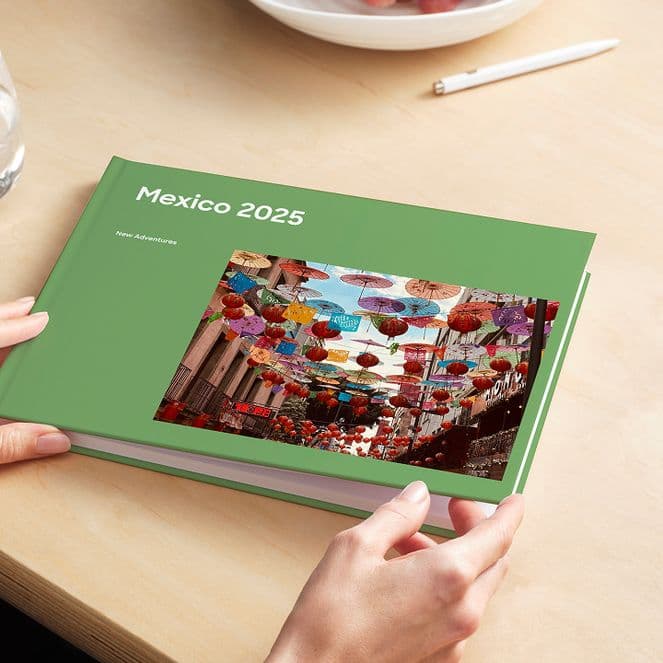 Person holding a green photo book titled "Mexico 2025" with a colorful market scene on the cover, on a wooden table with a glass of water.