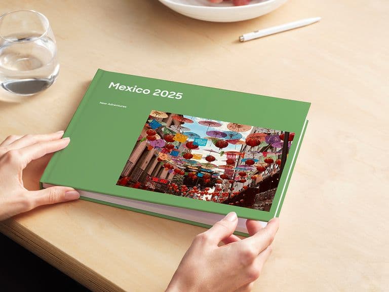 Person holding a green photo book titled "Mexico 2025" with a colorful market scene on the cover, on a wooden table with a glass of water.
