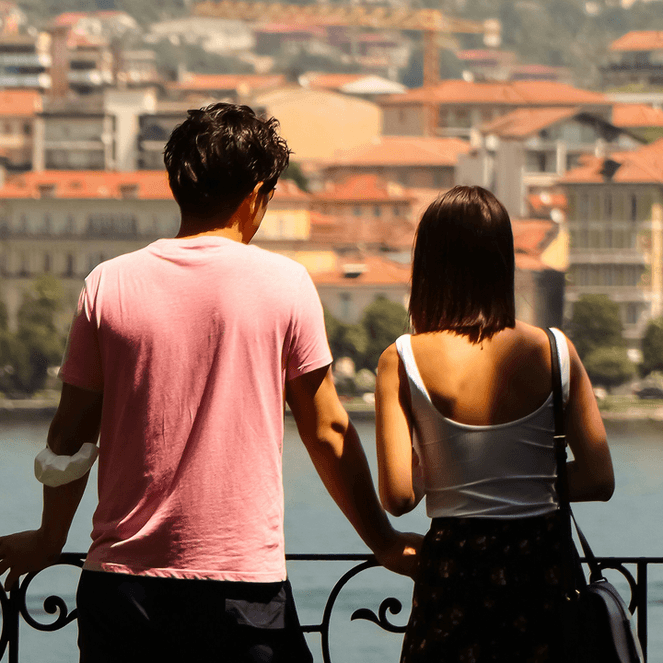 A couple leans on a railing, overlooking a scenic view of a lake and a town with red-roofed buildings in the background.