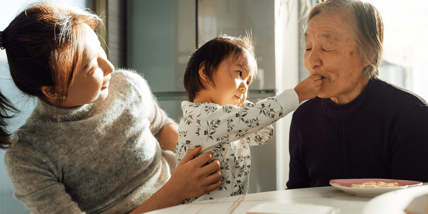 A child playfully feeds an elderly relative while her mother smiles, sitting together at a table.