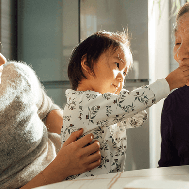 A child playfully feeds an elderly relative while her mother smiles, sitting together at a table.