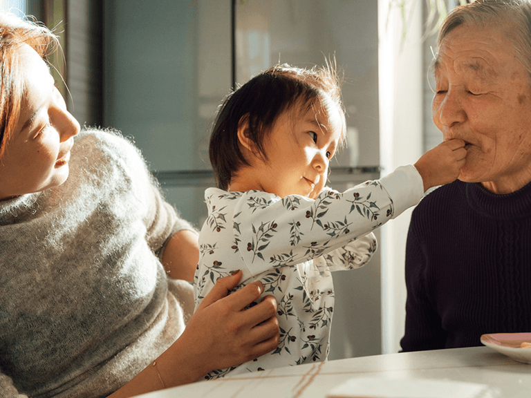 A child playfully feeds an elderly relative while her mother smiles, sitting together at a table.