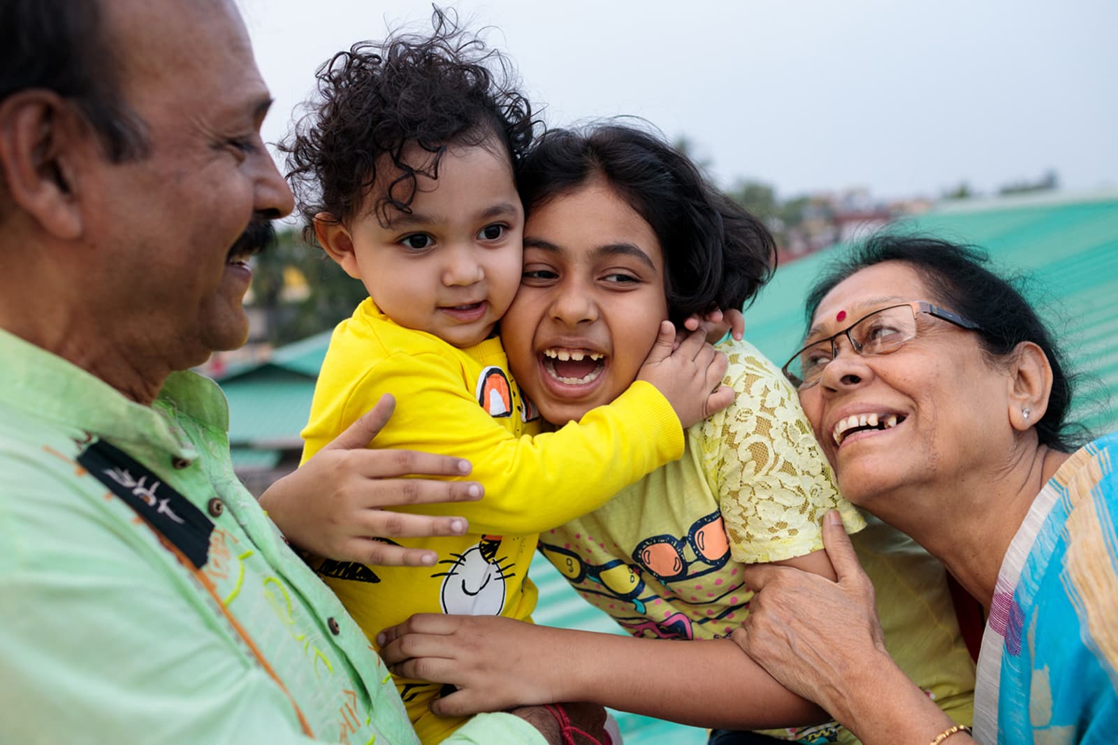 A joyful family moment with grandparents smiling at their two young grandchildren, who are hugging each other, outdoors on a bright day.