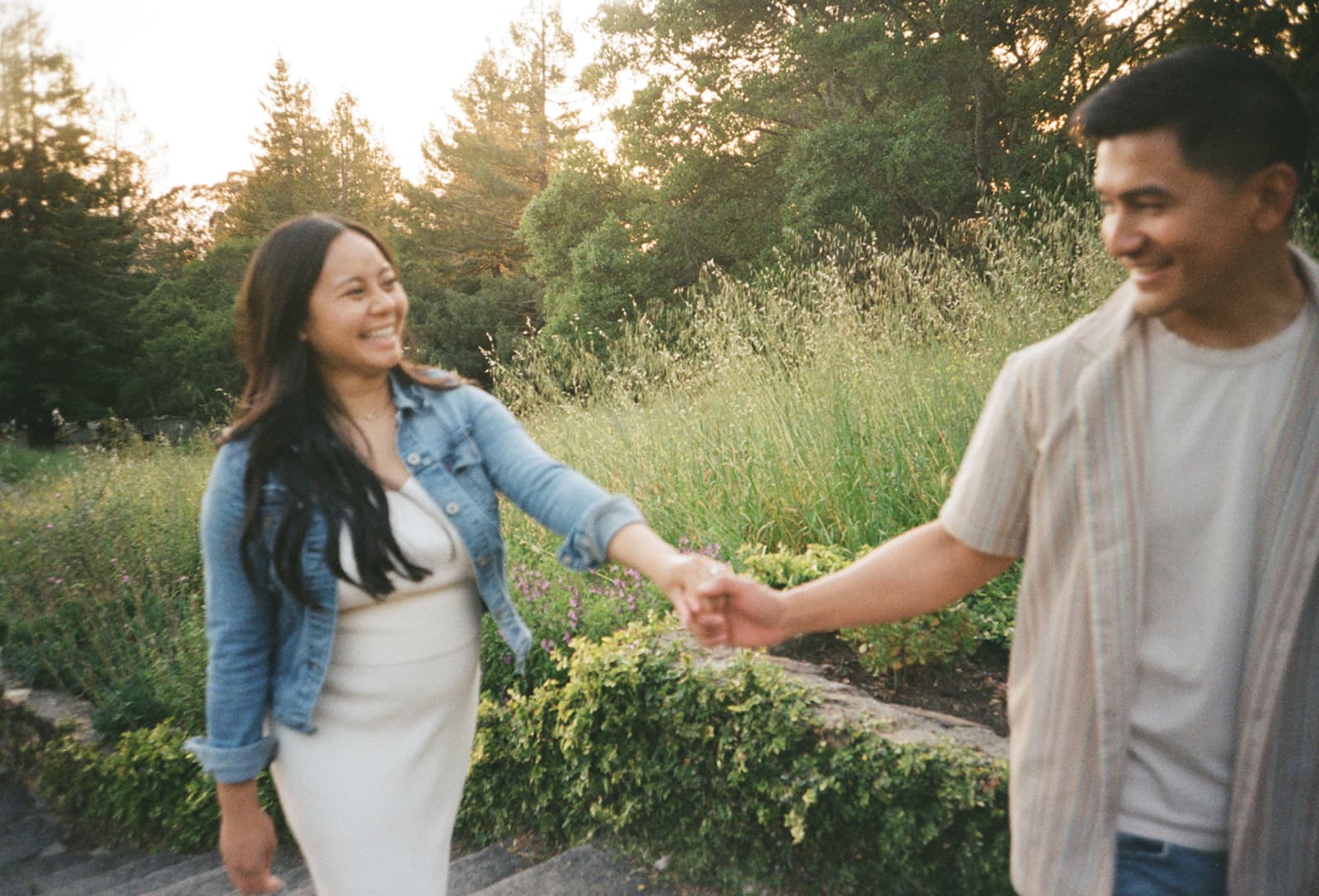 A couple smiling and holding hands while walking outdoors, surrounded by greenery and soft sunlight.