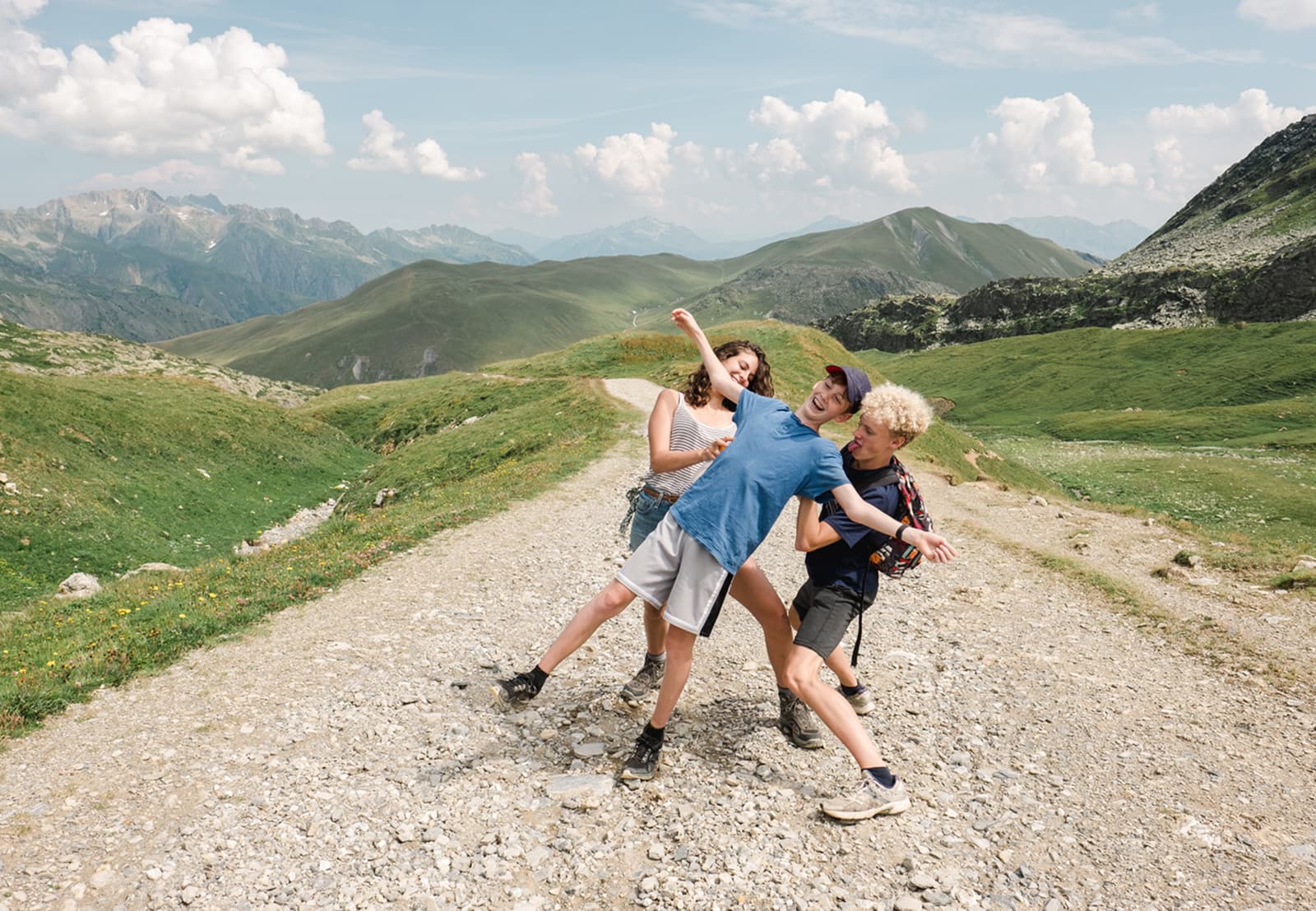 Four people joyfully pose on a rocky mountain path, surrounded by green hills and distant peaks under a blue sky with fluffy clouds.