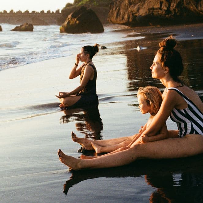Two women and a child sat meditating at the water's edge on a beach.