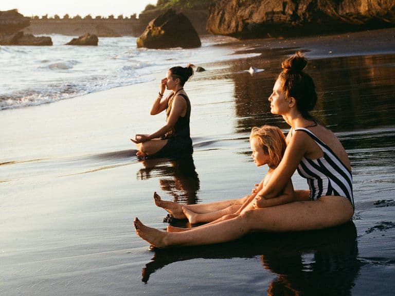 Two women and a child sat meditating at the water's edge on a beach.