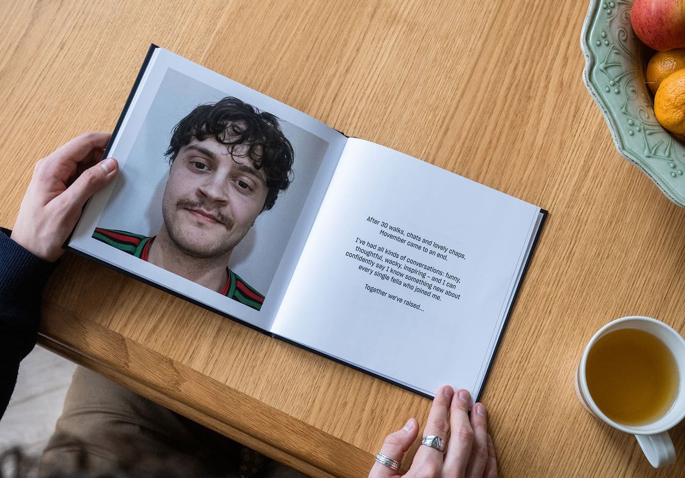 Person holding a photo book open to a page with a portrait on the left and text on the right, next to a cup and bowl of fruit on a wooden table.