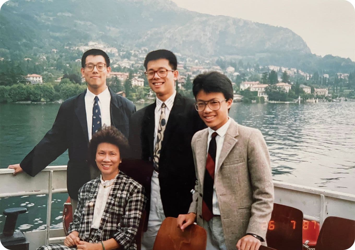 Four people posing on a boat with a scenic lake and mountains in the background, dressed in suits and smiling.