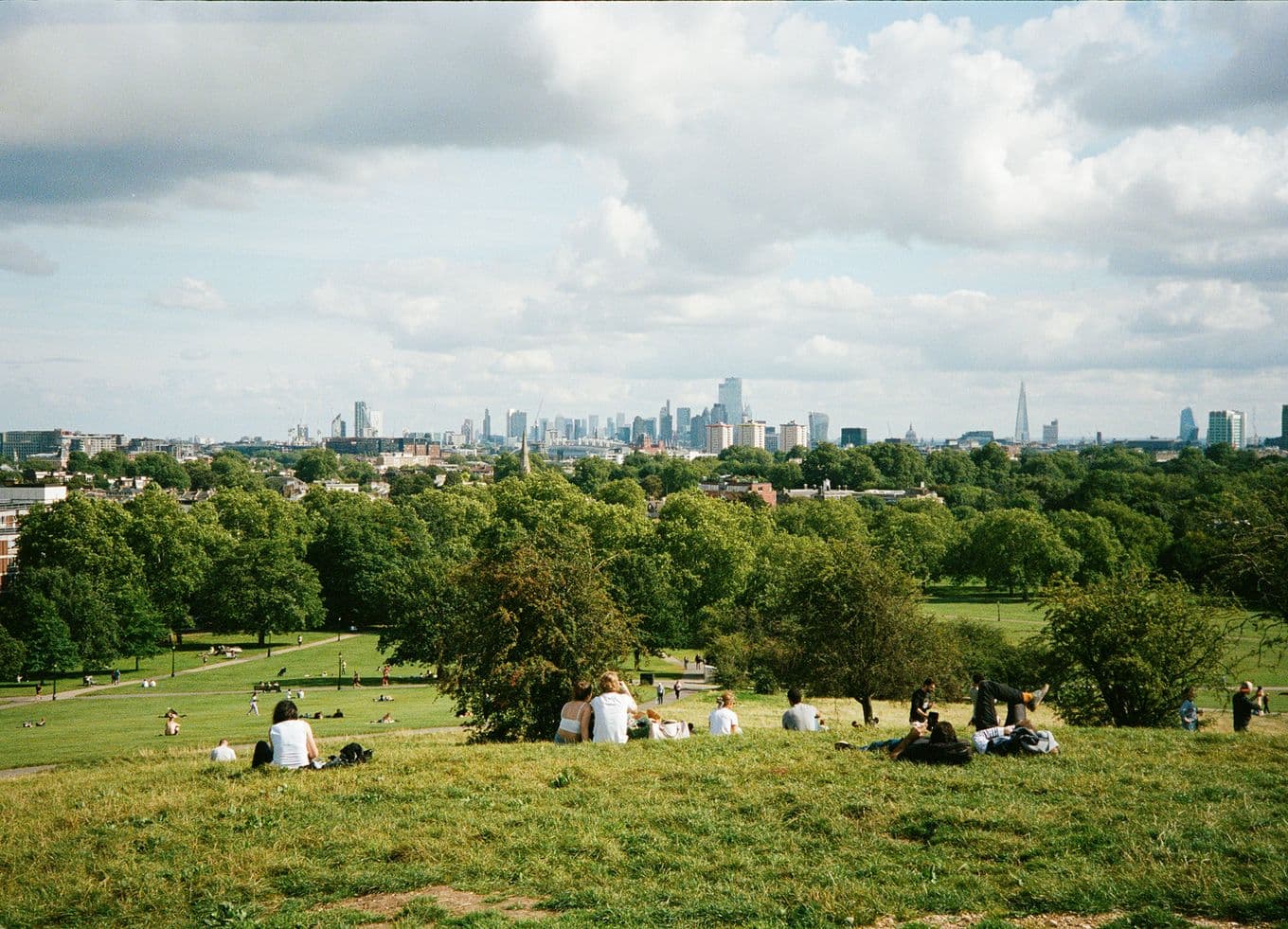 People relaxing on a grassy hill overlook a sprawling city skyline with patches of trees and cloudy skies in the background.